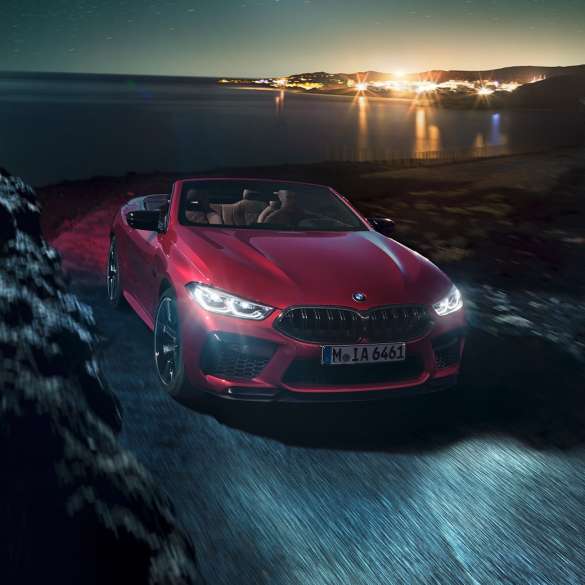 BMW M8 Competition Cabrio, kolm neljandikku eestvaade. BMW M8 Competition Cabrio toonis Motegi Red metallik, avatud pehme katusega, kolm neljandikku eestvaade, öösel rannikuteel sõitmas.