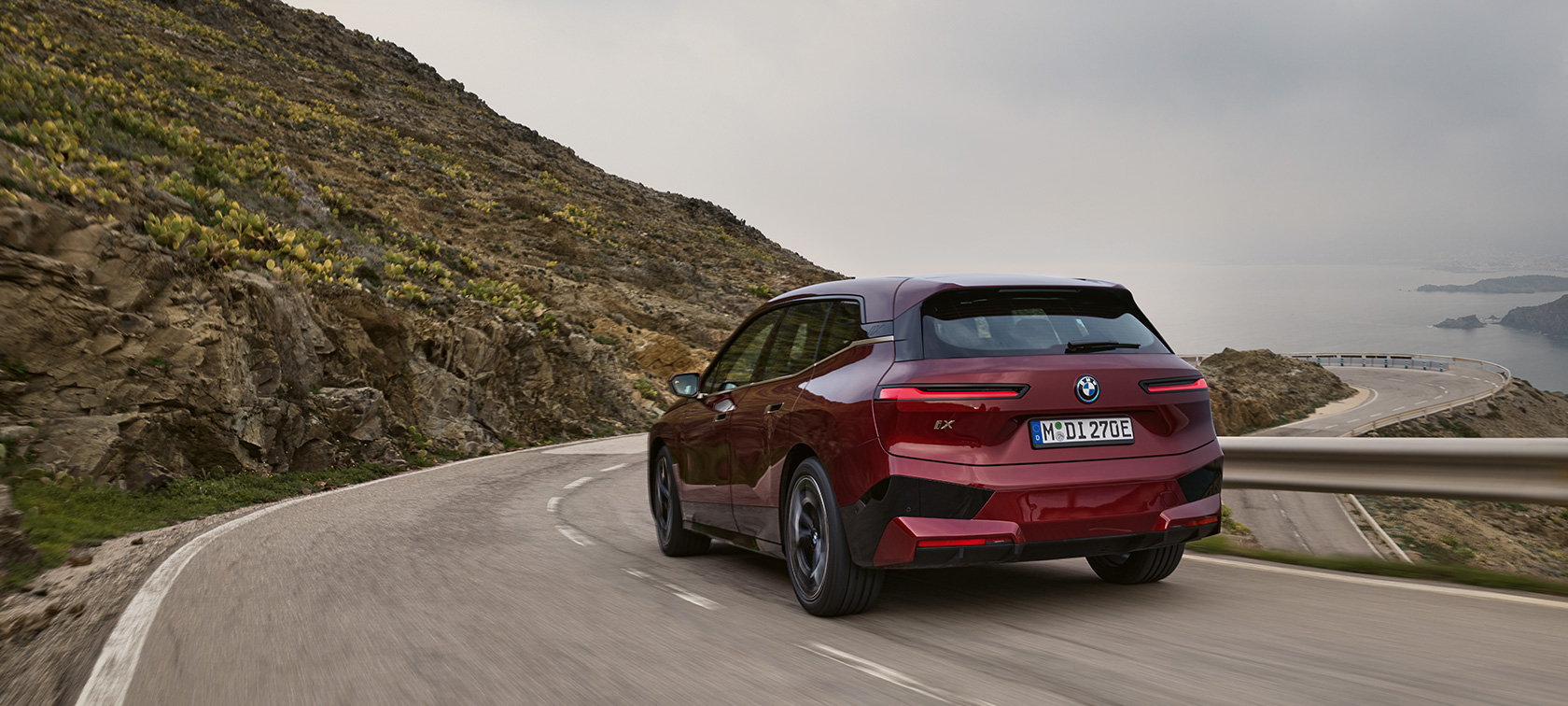 BMW iX i20 2021, elektriline SUV BMW iX xDrive50, sõitmas mereäärsel maanteel BMW iX i20 2021, elektriline SUV BMW iX xDrive50 Aventurin Red tagantvaates, sõitmas mereäärsel maateel