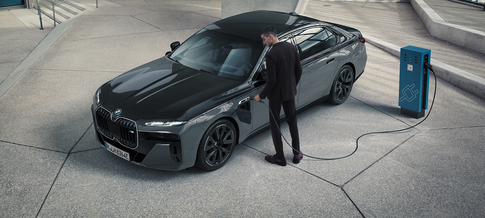 BMW M760e xDrive Sedaan G70, M Performance’i pakett, BMW Individuali kahetooniline värviviimistlus Black Sapphire metallik, BMW Individual Dravite Grey metallik, kolm neljandikku eestvaade, laadimiskaabliga mees laadimisjaama kõrval