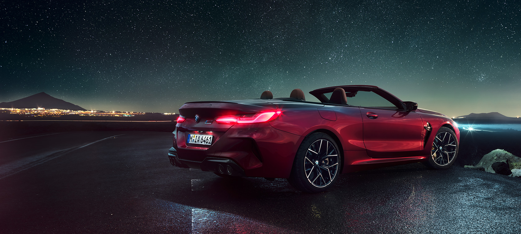 BMW M8 Competition Cabrio, kolm neljandikku tagantvaade. BMW M8 Competition Cabrio toonis Motegi Red metallik, avatud pehme katusega, kolm neljandikku tagantvaade, seismas tähistaeva all.