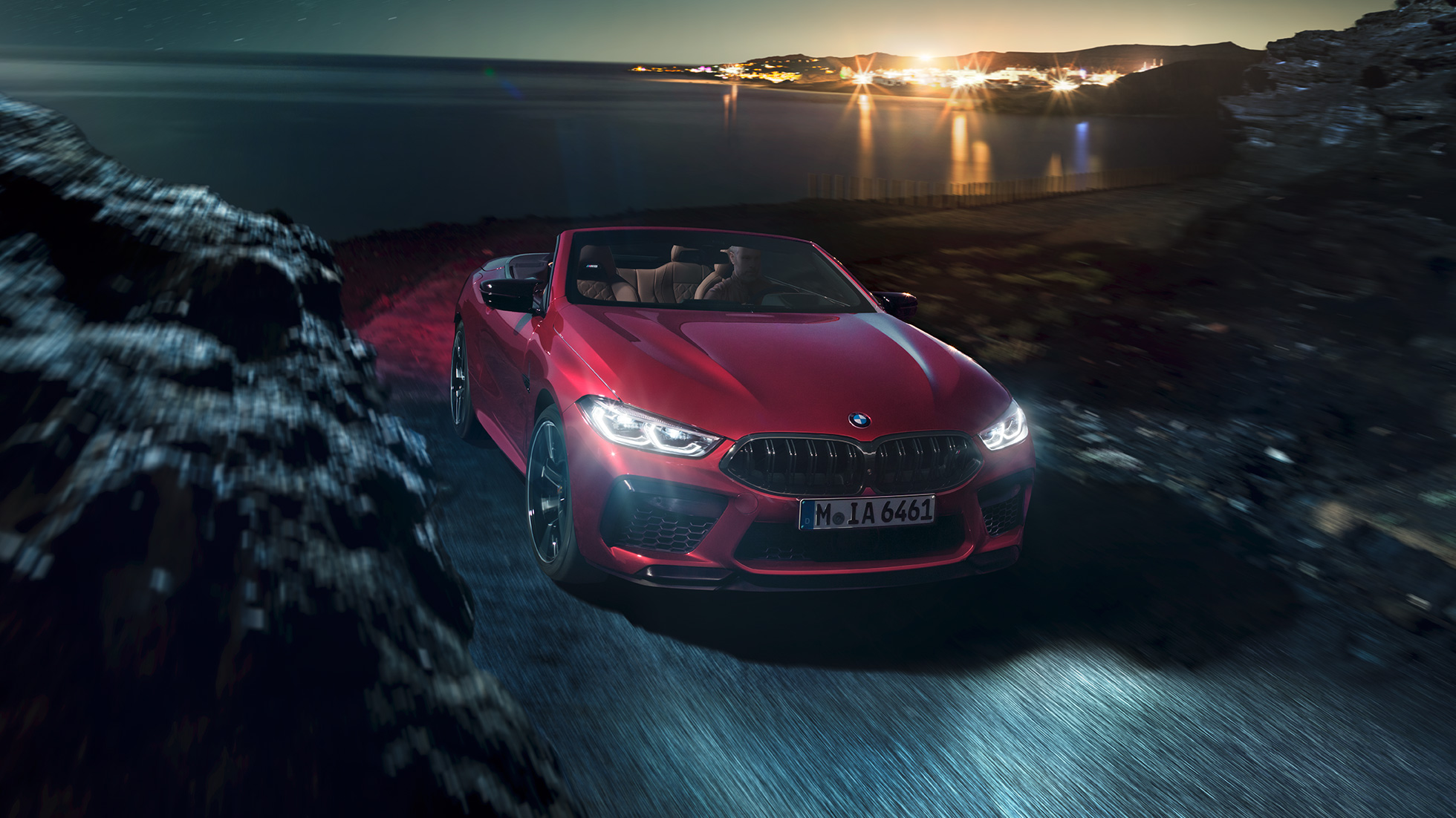 BMW M8 Competition Cabrio, kolm neljandikku eestvaade. BMW M8 Competition Cabrio toonis Motegi Red metallik, avatud pehme katusega, kolm neljandikku eestvaade, öösel rannikuteel sõitmas.