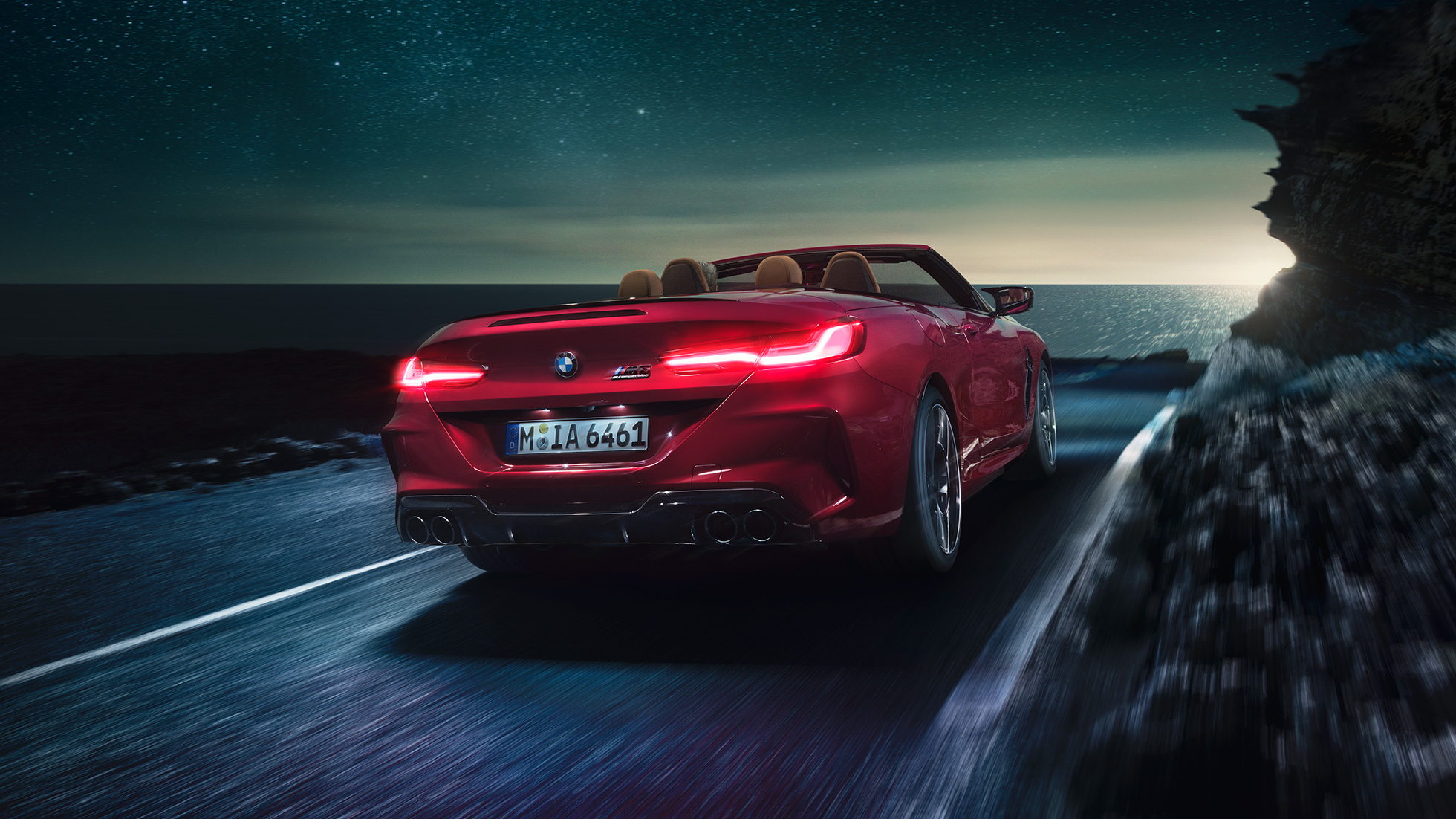 BMW M8 Competition Cabrio, kolm neljandikku tagantvaade. BMW M8 Competition Cabrio toonis Motegi Red metallik, avatud pehme katusega, kolm neljandikku tagantvaade, öösel rannikuteel sõitmas.