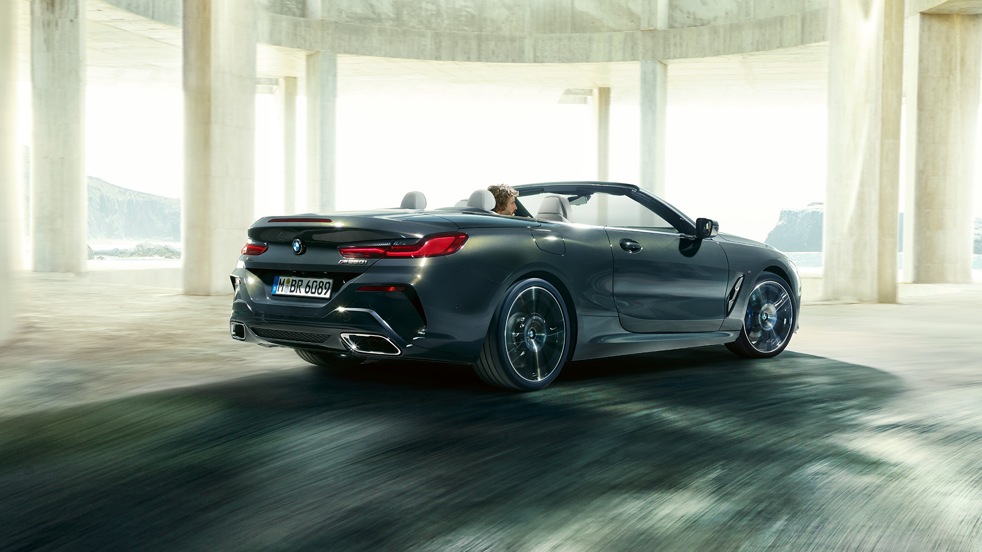 BMW M850i xDrive Cabrio, kolm neljandikku tagantvaade. BMW M850i xDrive Cabrio toonis Dravit Grey metallik, avatud pehme katusega, kolm neljandikku tagantvaade, sõitmas läbi betoonehitise.