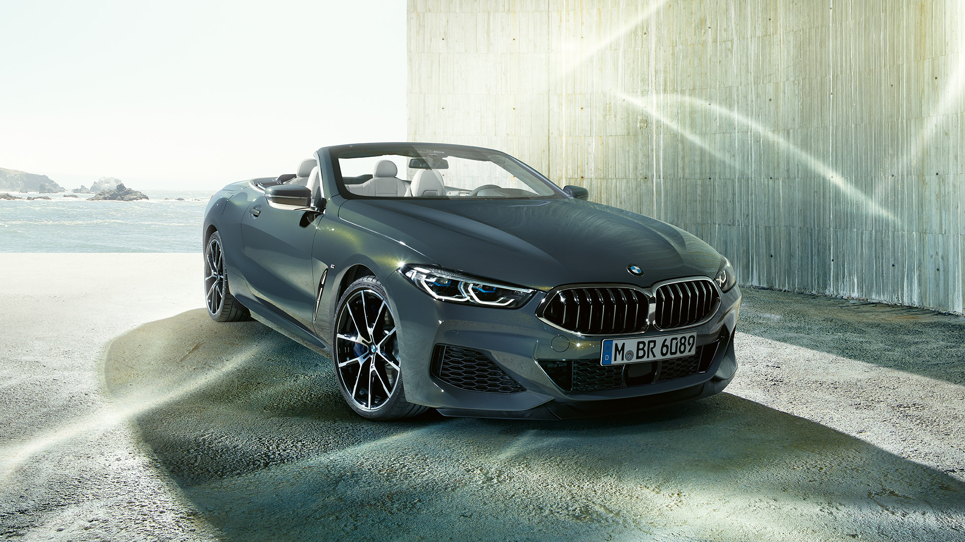 BMW M850i xDrive Cabrio, kolm neljandikku eestvaade. BMW M850i xDrive Cabrio toonis Dravit Grey metallik, avatud pehme katusega, kolm neljandikku eestvaade, seismas ranniku taustal.
