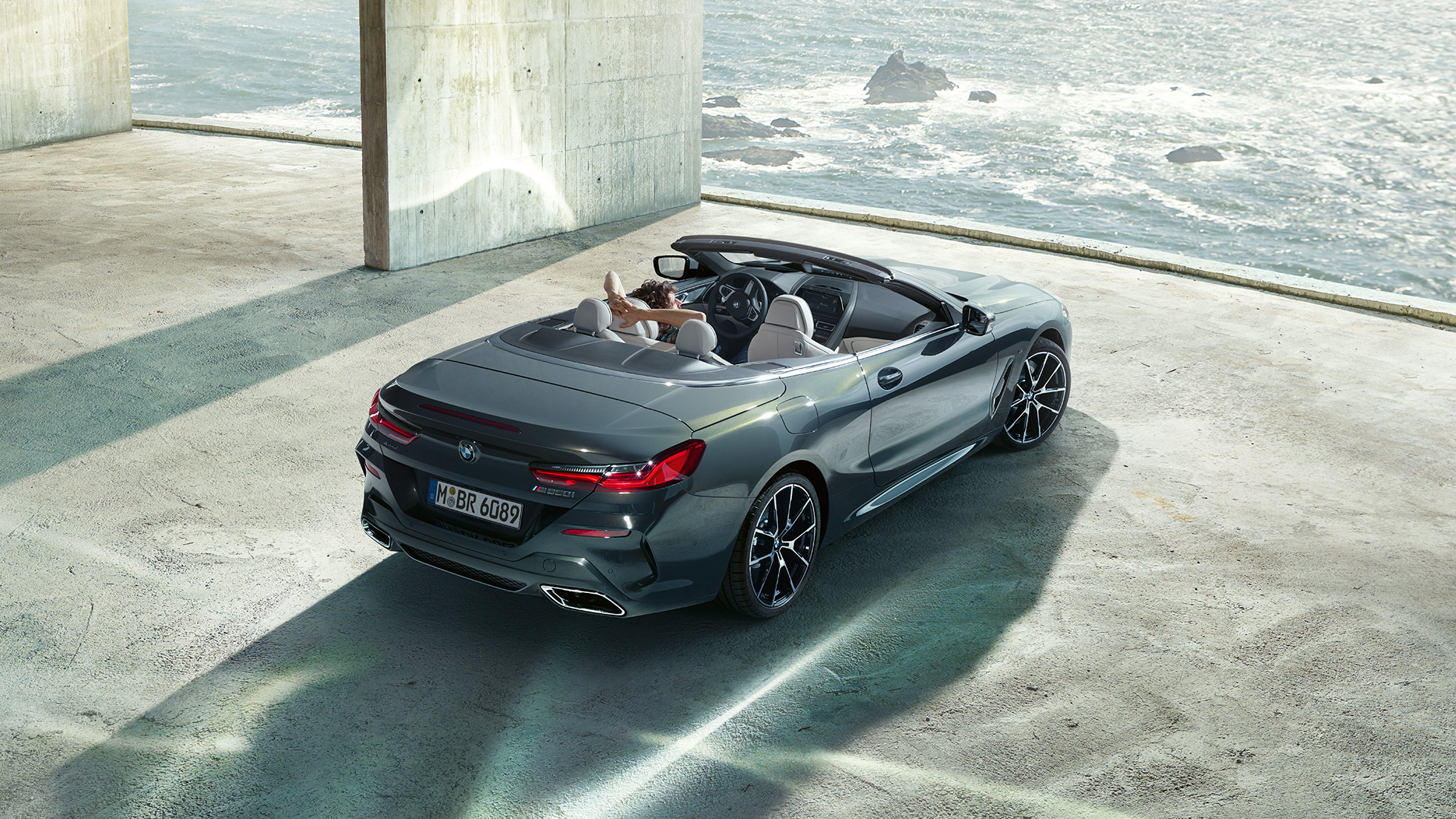 BMW M850i xDrive Cabrio, ülaltvaade. BMW M850i xDrive Cabrio toonis Dravit Grey metallik, avatud pehme katusega, ülaltvaade, parkimas betoonehitise ees ookeani taustal.