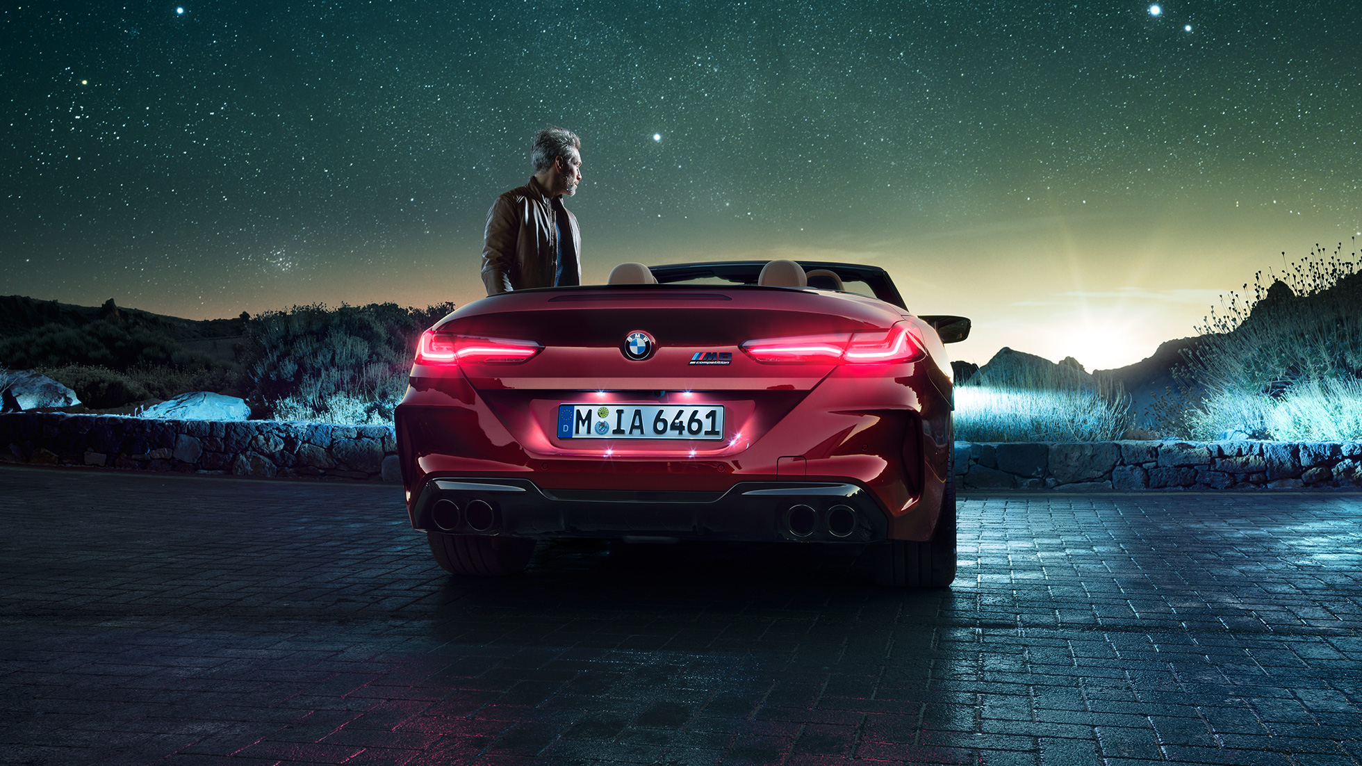 BMW M8 Competition Cabrio, tagantvaade. BMW M8 Competition Cabrio toonis Motegi Red metallik, avatud pehme katusega, tagantvaade, seismas koos mehega tähistaeva all.