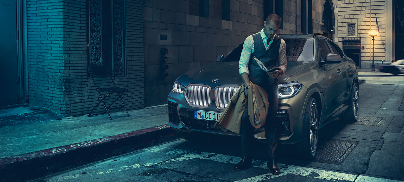Mees nõjatumas vastu BMW X6. Man leaning on BMW X6 in lateral perspective in front of an urban setting.