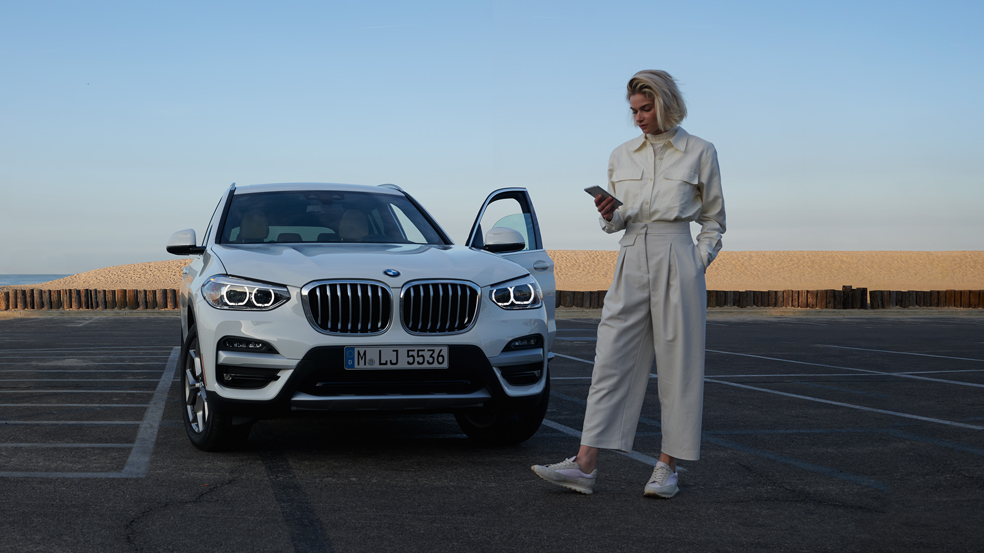 Naine kasutamas My BMW rakendust nutitelefonis Naine kasutamas BMW ConnectedDrive’i rakendust My BMW nutitelefonis valge BMW X5 ees